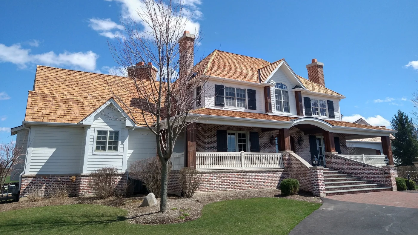 Remarkable Roofing Contractor | Bartlett, IL | Apex Exteriors, Inc. Traditional brick and siding home with a warm-toned cedar shake roof, multiple chimneys, and a wide covered front porch, showcasing detailed rooflines and classic window trim.