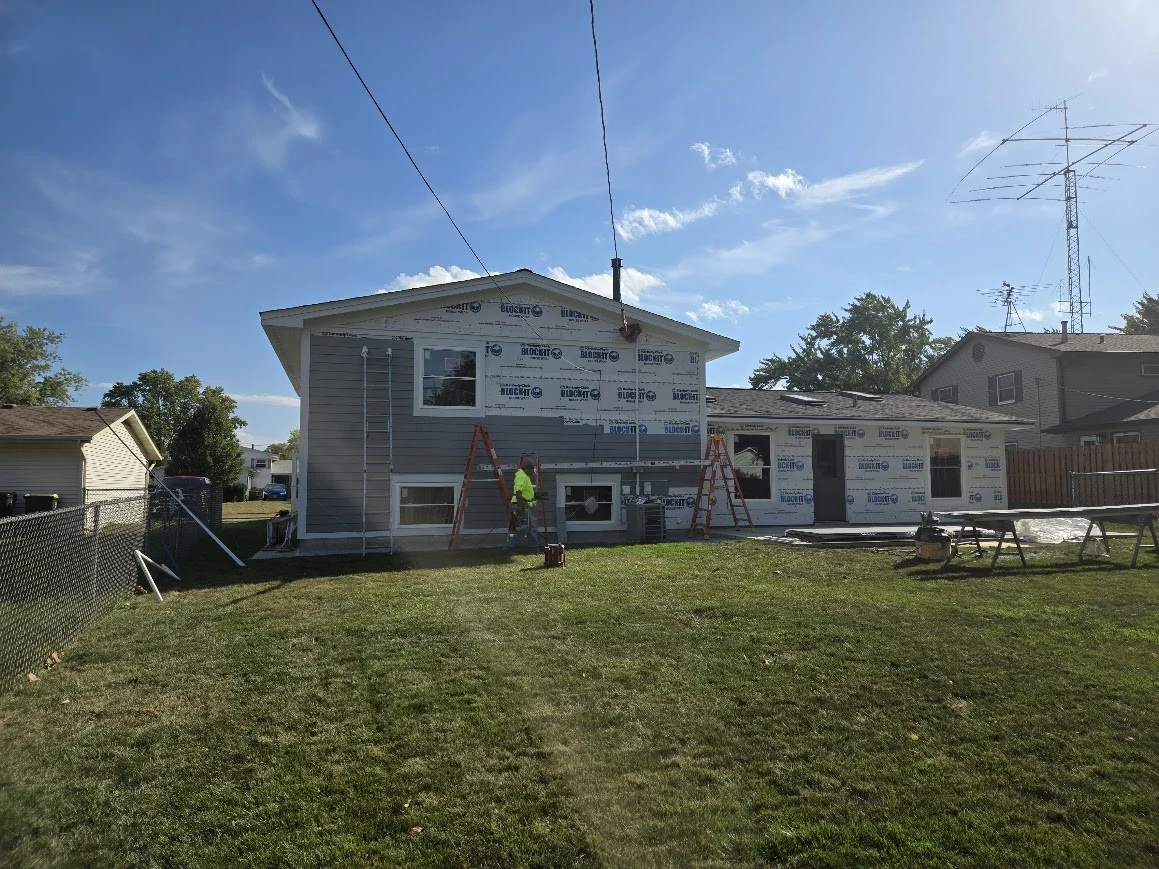 Residential siding installation in progress on a split-level home with house wrap exposed, ladders set up, and a worker in the yard under a sunny sky.