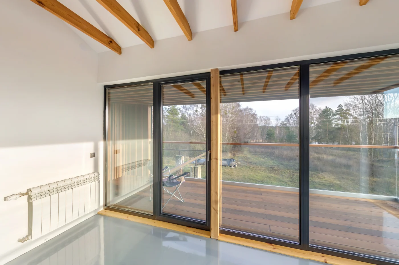 An interior view of a set of large windows and a sliding door to their left that leads out to a wooden deck and grassy backyard. The floors are light gray, the walls are white, and there are exposed wooden beams on the ceiling. The door and windows have black framing.