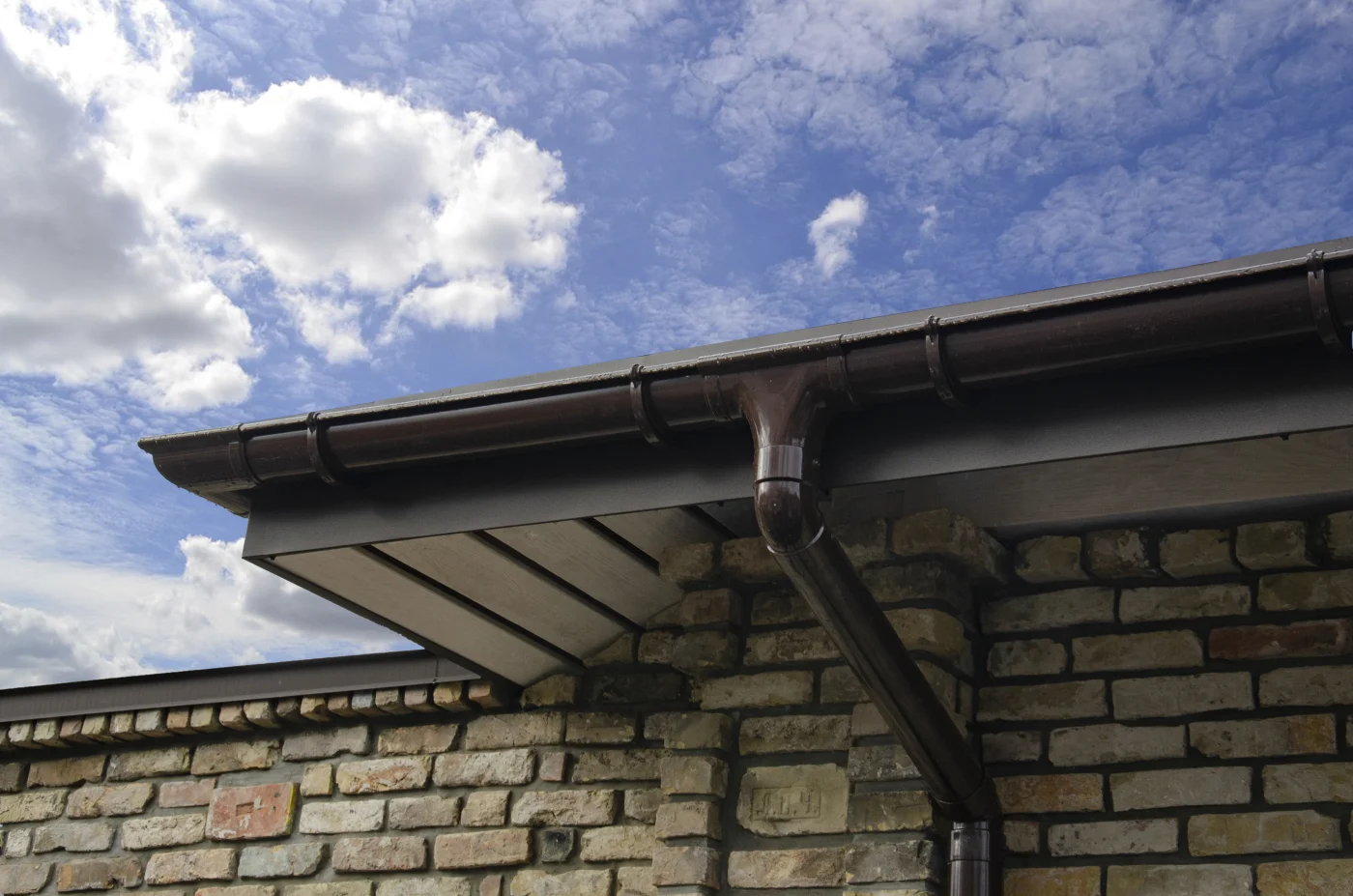 Sound Soffit And Fascia Replacement In Elgin, IL | Apex Exteriors The upper portion of a brick home has a dark brown gutter system. You can see a blue sky with puffy clouds above the home.