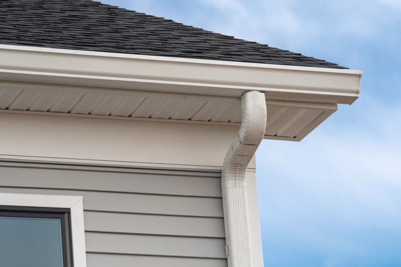 Standard Gutter Replacement In Elgin, IL |Apex Exteriors One corner of a home’s roof with the sky in the background. The home’s siding is light gray, the shingled roof is dark gray, and the gutters, fascia, and soffit are white. The right top corner of a window is visible just below the roofline.