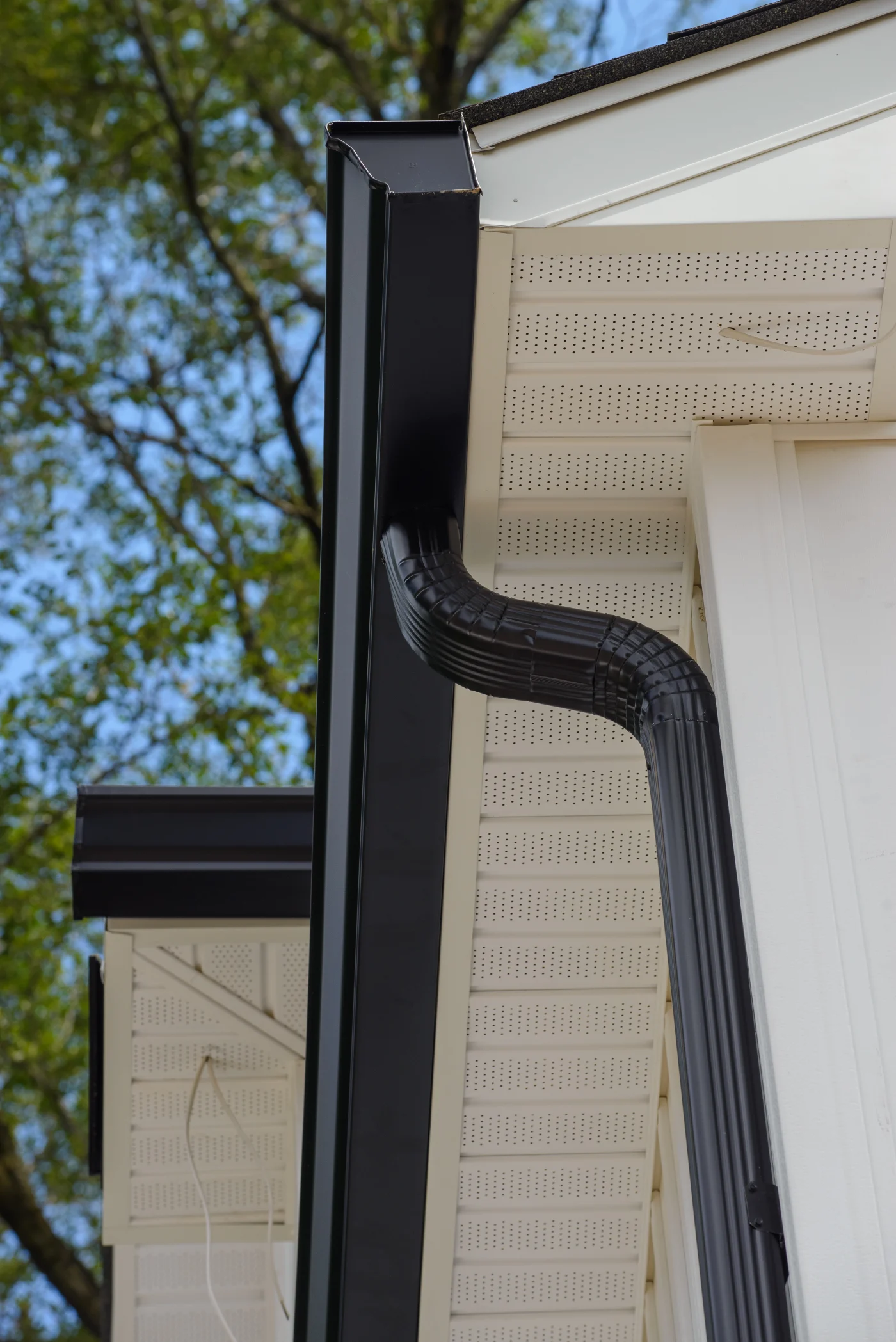 A view looking up at the underside of a home's gutter, soffit, and fascia. The home is white, and the gutter system is black. You can see green trees and the blue sky in the background.