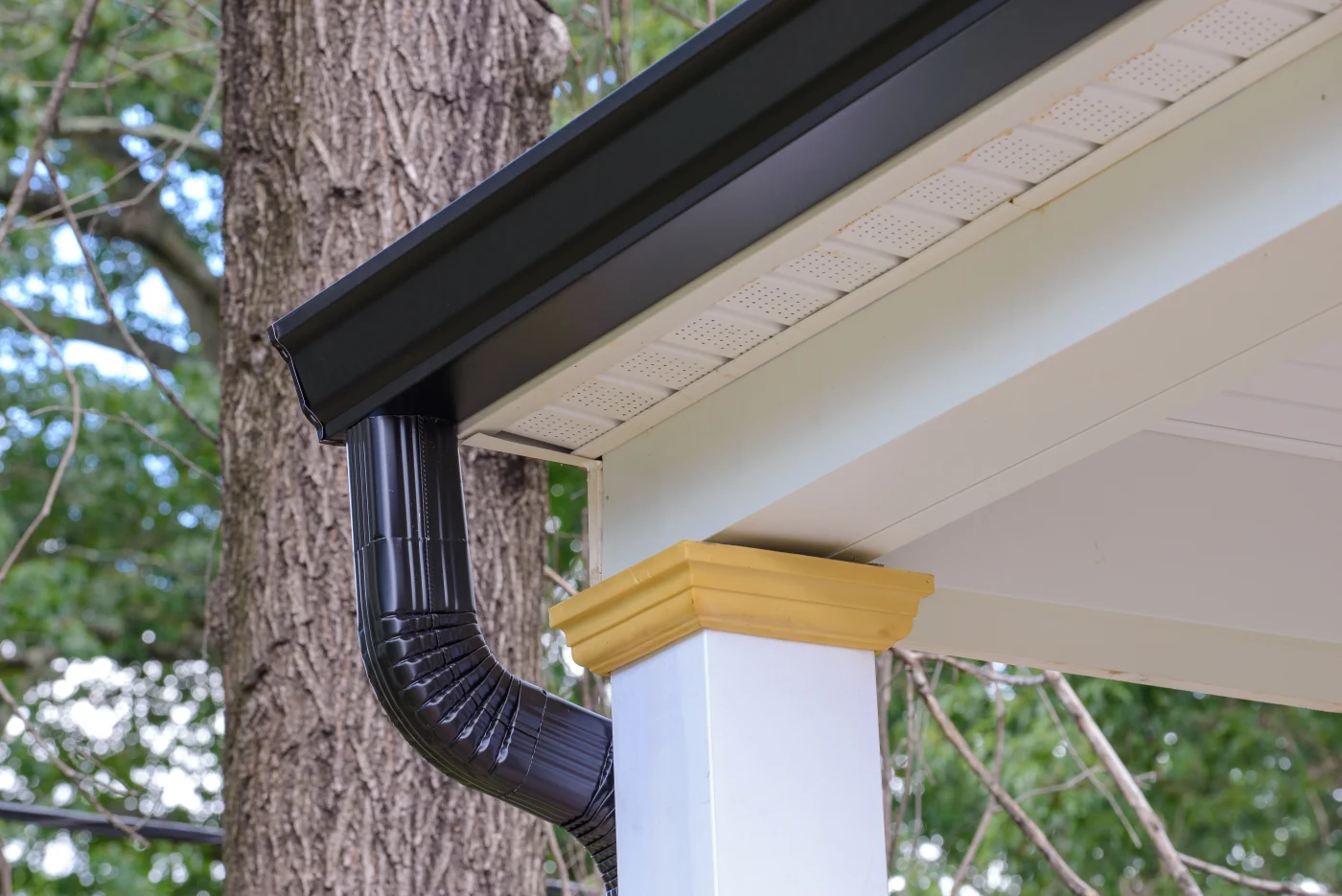 Looking up from the ground, you can see one corner of a home’s roof. The gutter and downspout are black, the soffit and fascia are white, and there is a white column support visible. In the background, there is a tree trunk with a few branches, and the sky is visible behind it.