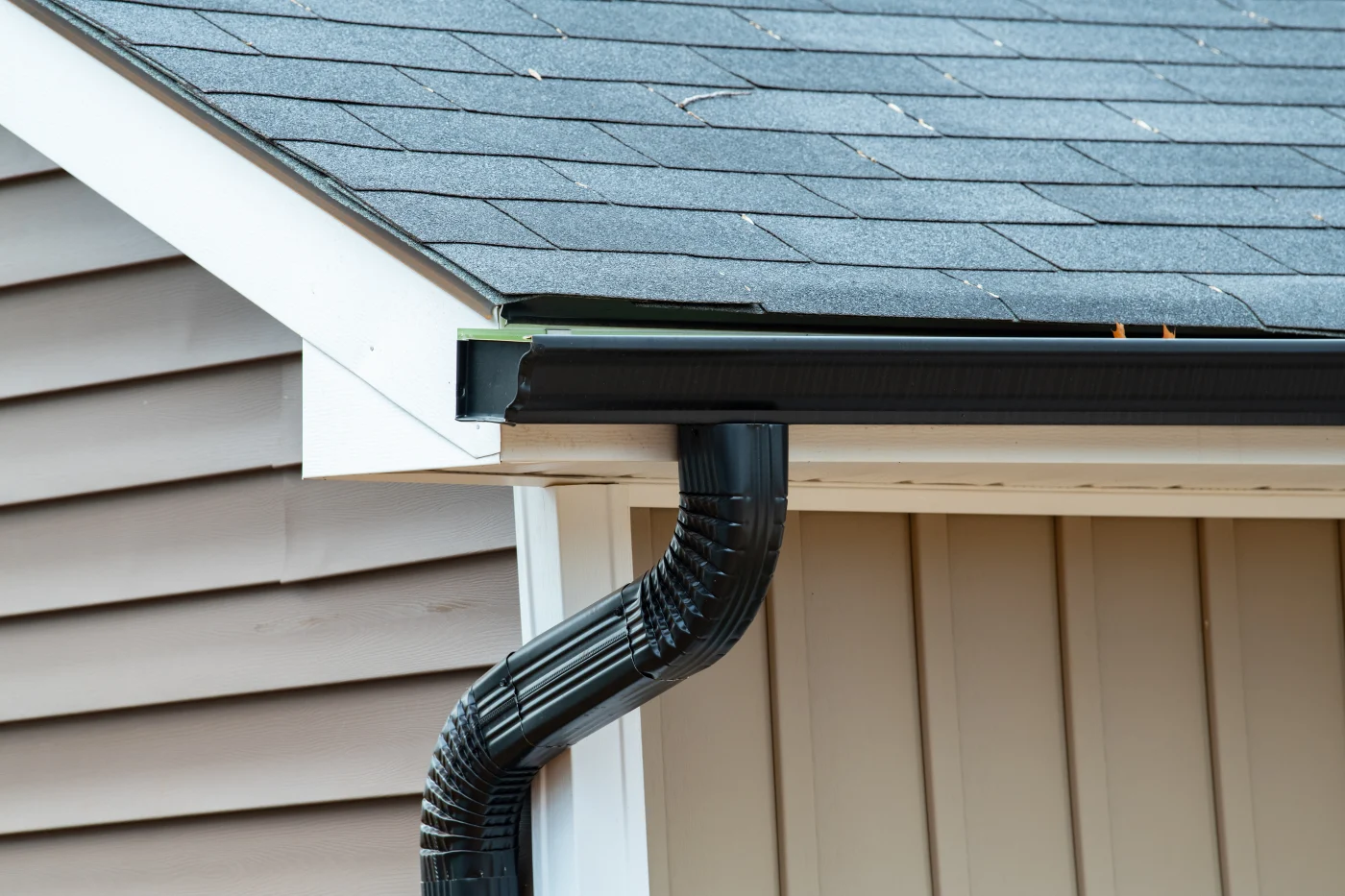 One corner of a home’s roof, showing tan siding, white trim, a dark gray roof, and black gutters and downspout.