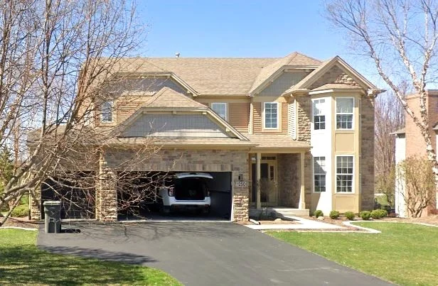 Trusted Roof Insurance Claims Assistance | Elgin, IL | Apex Exteriors A large two-story tan suburban home with a stone and siding exterior, an attached two-car garage with a white vehicle inside, and a blacktop driveway.