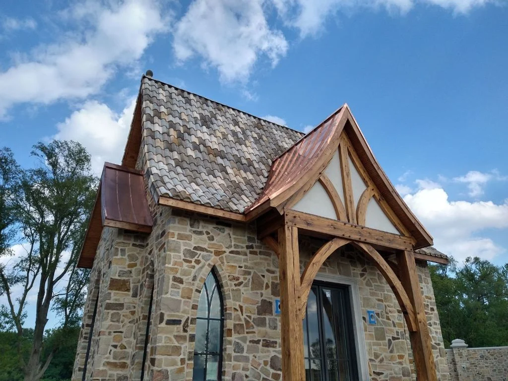 A rustic stone home with a steep gray tile roof, copper metal roof accents, timber-framed front entry, arched windows, and natural wood trim.