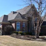 Two-story brick-and-stone home with multiple rooflines, arched windows, light-colored siding accents, and a landscaped front yard beneath a clear blue sky.